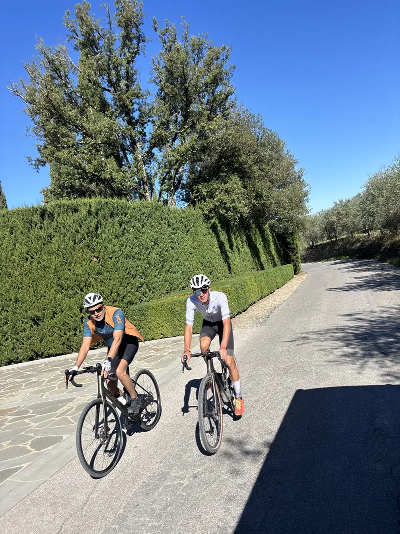 Two riders on a road beside a tall clipped hedge