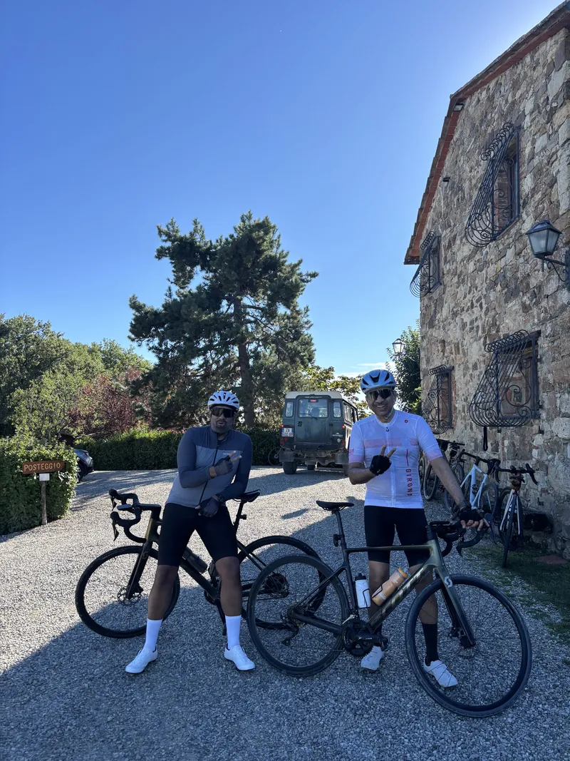 Two riders with their bikes outside an old stone house, the support van behind