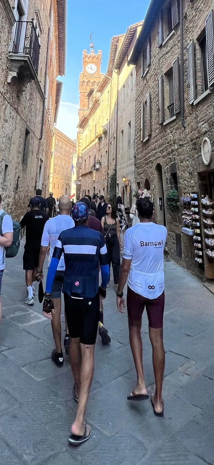 Riders in cycling kit walking along a narrow street towards a clock tower