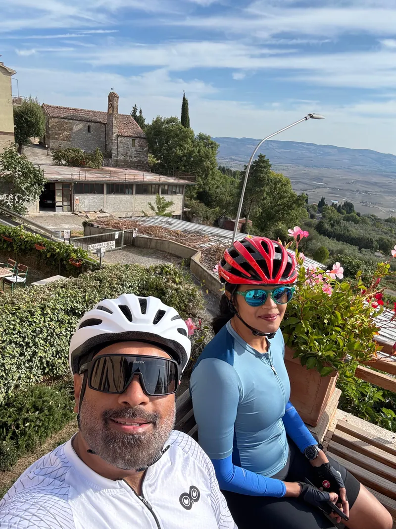 Two riders take a selfie on a terrace, a stone farmhouse and hills behind