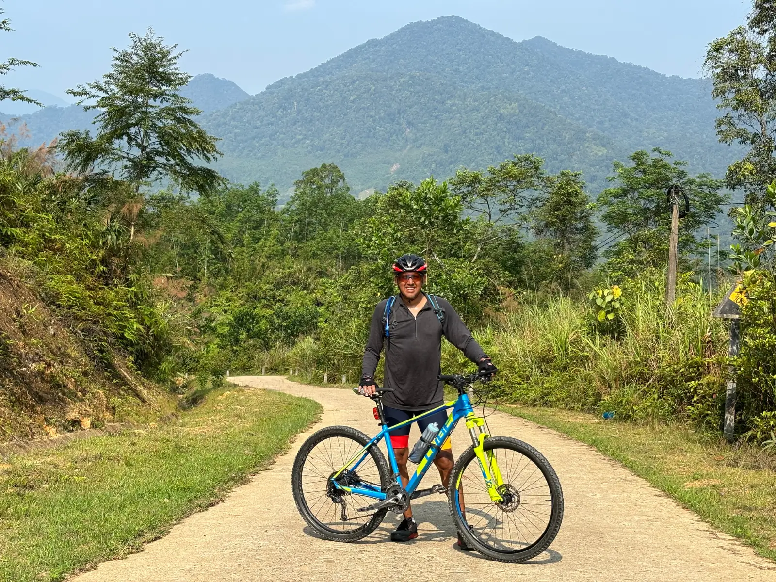 A rider standing with a mountain bike on a narrow concrete road in Vietnam, a forested peak behind